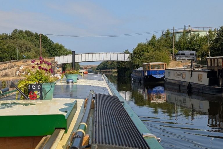 Narrowboat cruising on the Aire & Calder Navigation with a white bridge in the distance beneath a blue sky.